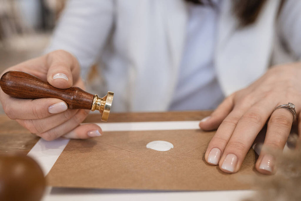 Close-up of hands with rings of unrecognizable woman making white wax seal with stamp on brown greeting invitation postcard envelope on white sheet of paper on wooden desk. Handmade craft, hobby. - Fotó, kép