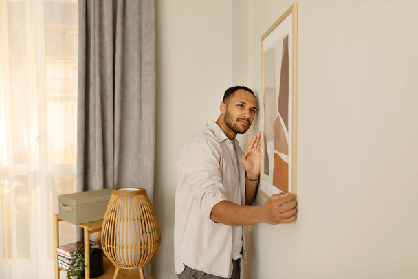 Portrait of handsome young African American man hanging picture frame on white wall at home, decorating his apartment, empty space. Modern interior design and home decor concept - Φωτογραφία, εικόνα
