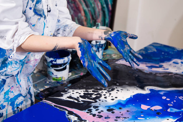 Detailní záběr na ruce umělce pokryté modrou barvou používanou jako nástroj k malování na plátně, na sobě bílý kabát pokrytý barevnými skvrnami - Fotografie, Obrázek