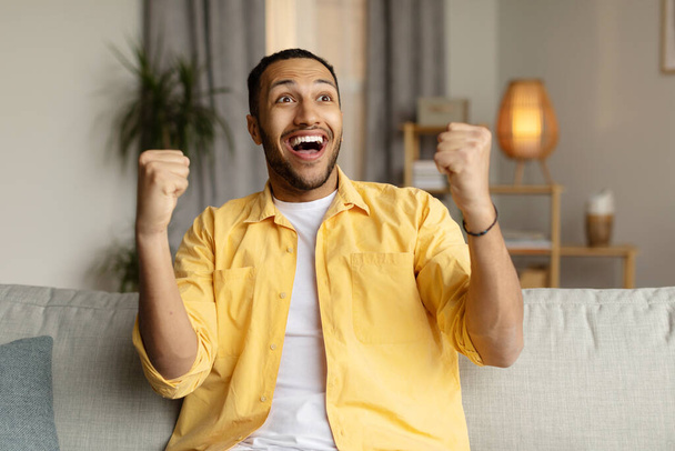 Boldog fiatal fekete férfi sikoltozik az izgalomtól, gesztikulál IGEN, otthon ül a kanapén. Diadalmas afro-amerikai férfi kiabál WOW, ünnepli a sikert, éljenzés a sport csapat a TV-ben - Fotó, kép