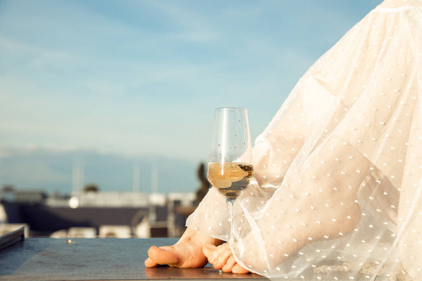 Cropped photo of elegant barefoot legs of woman wearing long light white transparent summer dress, holding glass of champagne with feet, sitting on edge of roof on background of blue sky and city. - Foto, afbeelding