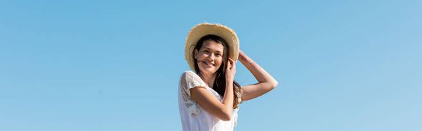 Χαμηλή γωνία άποψη της όμορφης γυναίκας στο καπέλο ήλιο χαμογελώντας στην κάμερα με μπλε ουρανό στο παρασκήνιο, πανό  - Φωτογραφία, εικόνα