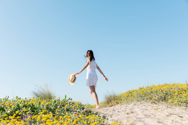 Jovem mulher segurando chapéu de sol enquanto corre na praia  - Foto, Imagem