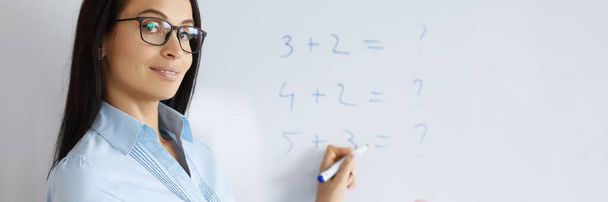 Portrait of young teacher at school explain maths to pupils. Examples with numbers on board, qualified professor. Copy space in right. Education, knowledge, school, university concept - Photo, Image