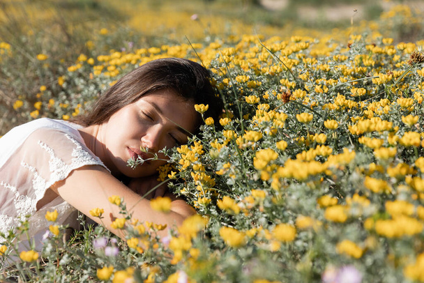 Брюнетка с закрытыми глазами лежит на желтых цветах на газоне  - Фото, изображение