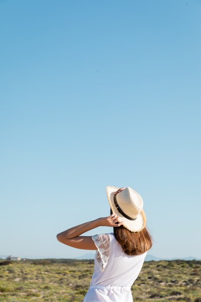 Vista posterior de la mujer morena en vestido blanco y sombrero de sol de pie en la playa borrosa  - Foto, Imagen