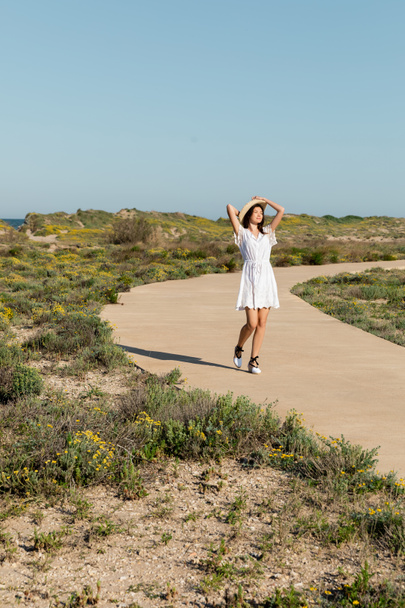 Young woman in summer dress and sun hat walking on pathway on beach  - Fotoğraf, Görsel