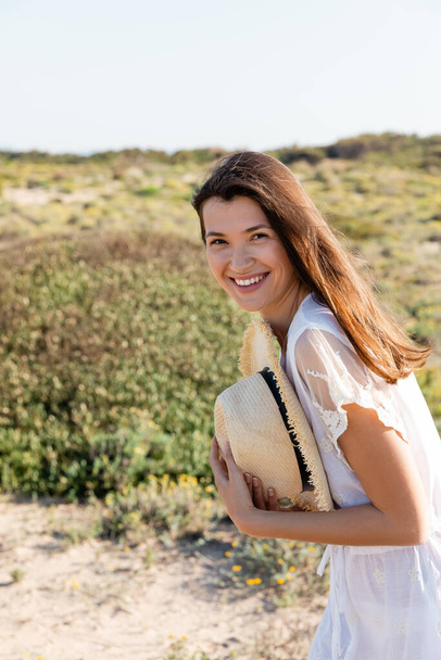 Pozitív nő nyári ruhában szalmakalapot tart a homályos strandon  - Fotó, kép