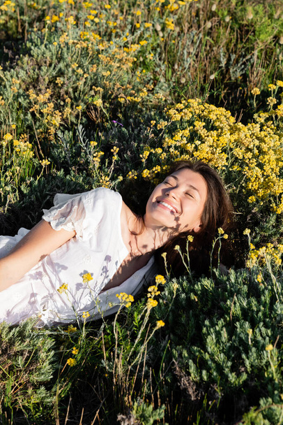 Cheerful woman in white dress lying on lawn with yellow flowers  - Fotó, kép