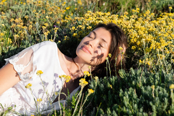 Brunette woman in white dress lying near flowers and grass on meadow  - Фото, изображение