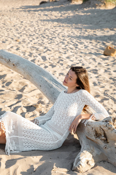 Pretty young woman in knitted dress sitting on sand near wooden log on beach  - Foto, Imagem