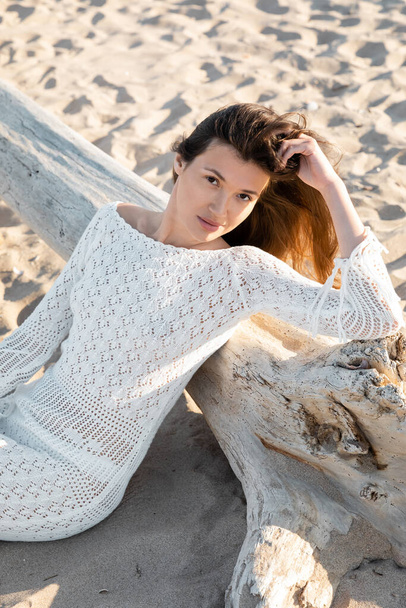 Vue grand angle de la jeune femme en robe tricotée d'été regardant la caméra près de la bûche sur la plage  - Photo, image