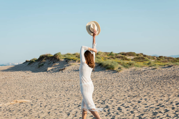 Vista trasera de la mujer en vestido de punto sosteniendo sombrero de sol en la playa de arena  - Foto, Imagen