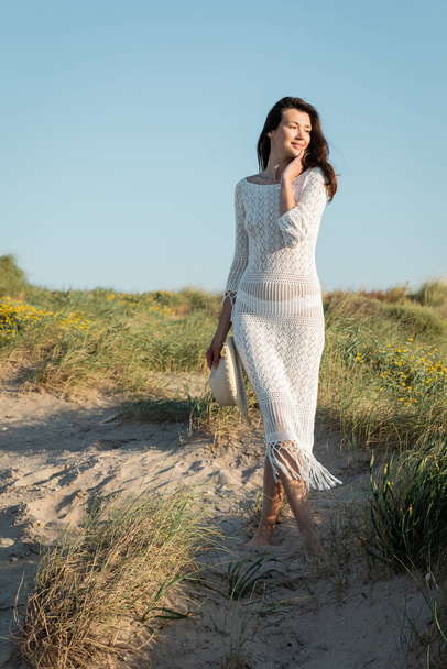 Mujer descalza en vestido de punto sosteniendo sombrero de sol mientras está de pie en la playa  - Foto, Imagen