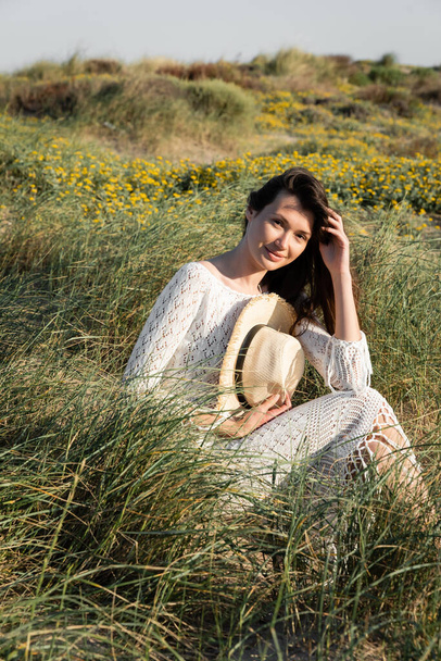 Mujer morena en vestido de verano sosteniendo sombrero de paja mientras está sentado en la hierba en la playa  - Foto, imagen