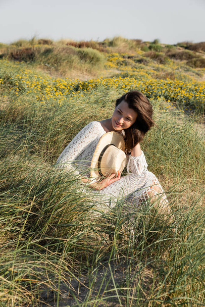 Uśmiechnięta kobieta trzyma kapelusz słoneczny siedząc na trawie na plaży  - Zdjęcie, obraz