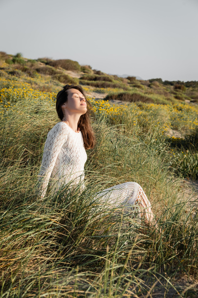 Brunette nainen sulkee silmänsä istuessaan ruohoa rannalla  - Valokuva, kuva