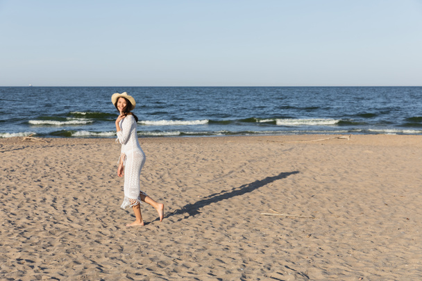 Positive woman in straw hat and summer dress walking near sea on beach  - Fotó, kép