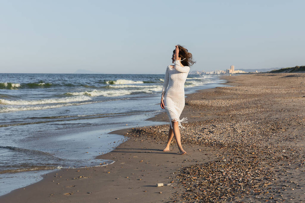 Вид сбоку улыбающейся женщины в вязаном летнем платье, прогуливающейся у моря  - Фото, изображение