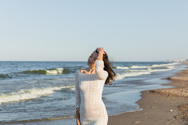 Brunette woman in knitted dress touching hair near sea on beach  - Zdjęcie, obraz