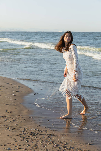 Positive young woman in knitted dress standing on wet sand on beach  - Fotografie, Obrázek