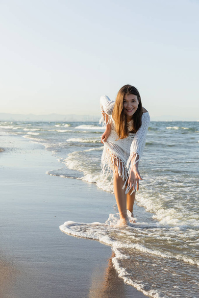 Happy woman in dress looking at camera while standing in sea water on sandy beach  - 写真・画像