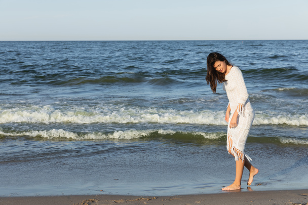 Πλήρες μήκος της μελαχρινής γυναίκας στο φόρεμα περπάτημα στην άμμο κοντά στη θάλασσα  - Φωτογραφία, εικόνα