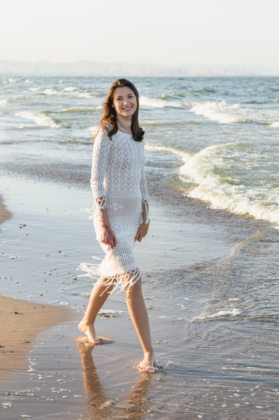 Χαμογελαστή γυναίκα με πλεκτό φόρεμα κοιτάζοντας κάμερα στην αμμώδη παραλία  - Φωτογραφία, εικόνα