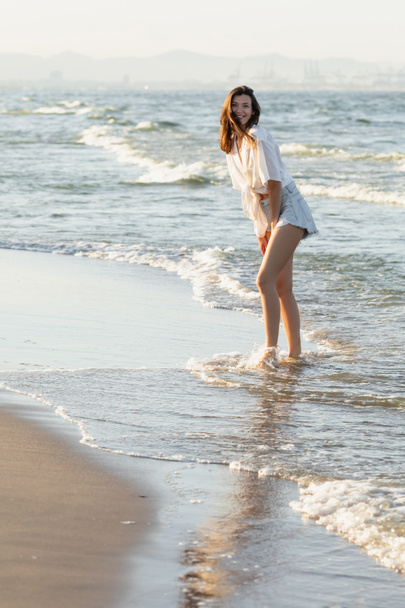 Femme souriante en chemise regardant la caméra tout en se tenant dans la mer  - Photo, image
