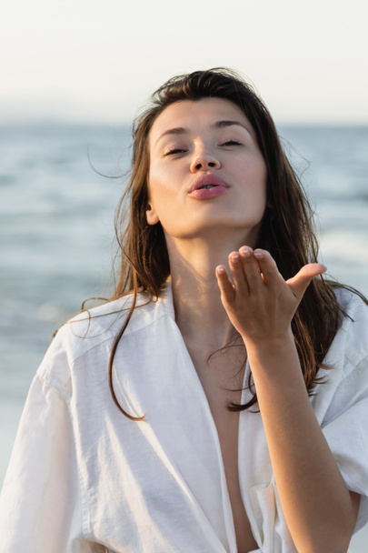 Morena mujer en camisa soplando aire beso cerca borrosa mar  - Foto, imagen