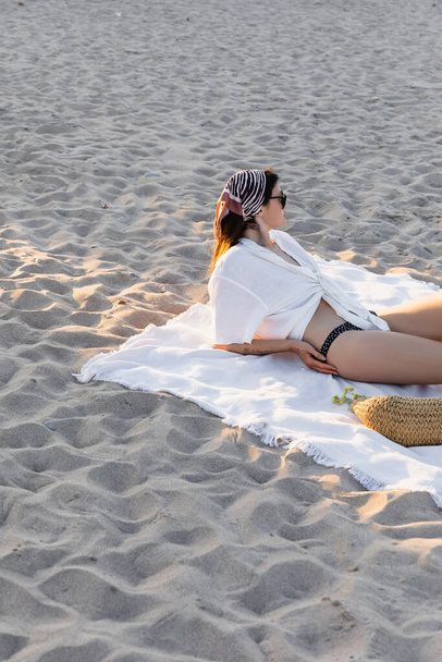 Side view of young woman in swimsuit and shirt lying on handbag on beach  - Foto, afbeelding
