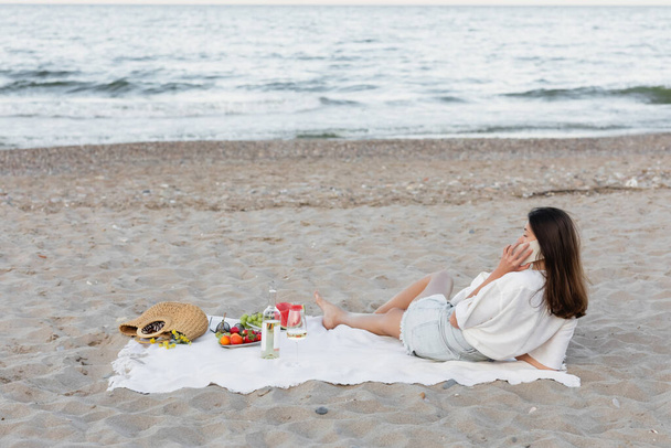 Junge Frau spricht auf Smartphone in der Nähe von Wein und Obst auf Decke am Strand  - Foto, Bild