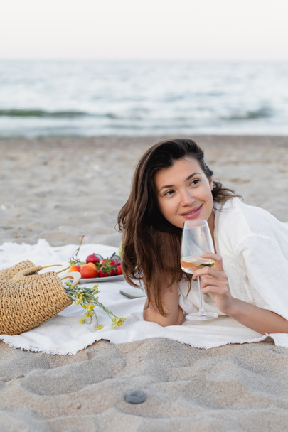 Mujer sonriente sosteniendo vino cerca del bolso con flores y frutas borrosas en la playa  - Foto, imagen