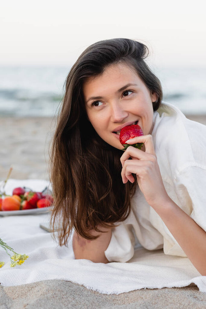 Uśmiechnięta brunetka jedząca truskawki na kocu na plaży  - Zdjęcie, obraz