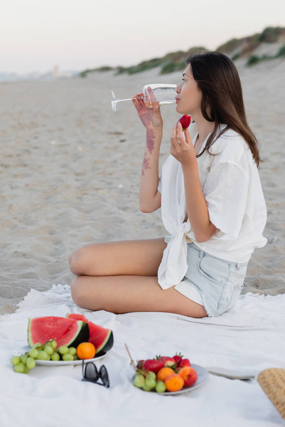 Side view of woman drinking wine and holding strawberry near blurred smartphone and fruits on beach  - Photo, Image