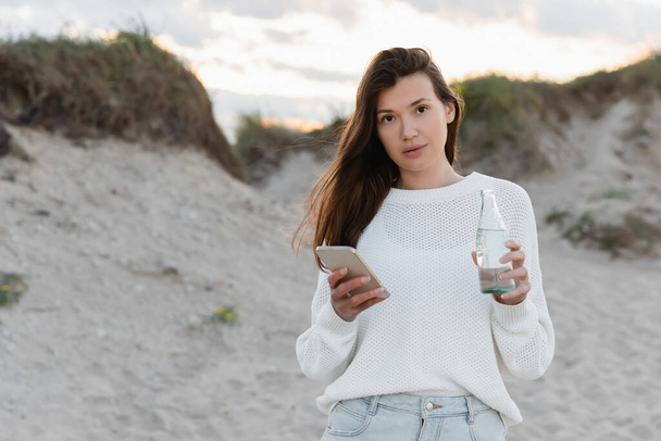 Brunette woman holding smartphone and bottle of water on beach in evening  - Foto, Imagen