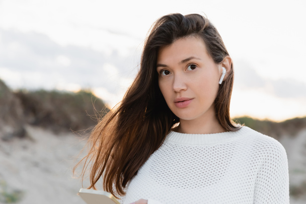 Brunette woman in earphone holding smartphone and looking at camera on beach  - Foto, Imagem