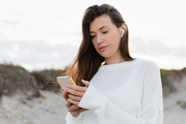 Young woman in earphone using cellphone on beach in evening  - Φωτογραφία, εικόνα