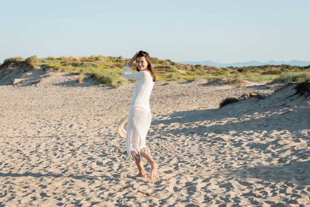 Mosolygós nő ruhában, napközben a strandon tartja a sapkáját.  - Fotó, kép