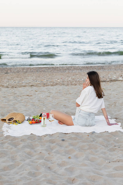 Donna che parla su smartphone vicino a vino e frutta sulla costa del mare  - Foto, immagini