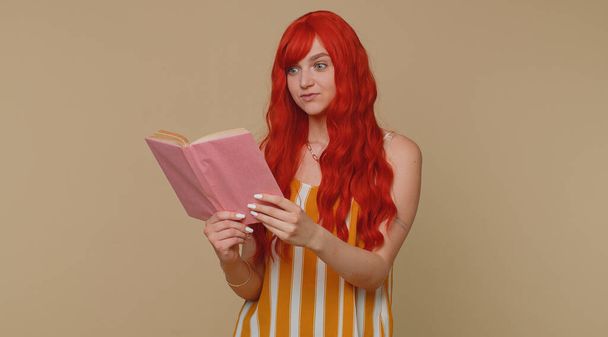 Рыжая молодая женщина читает забавную интересную сказку, увлекается досугом, мудростью знаний, образованием, обучением, учебой, вау. Рыжая рыжая девушка изолирована в одиночестве на фоне бежевой студии - Фото, изображение
