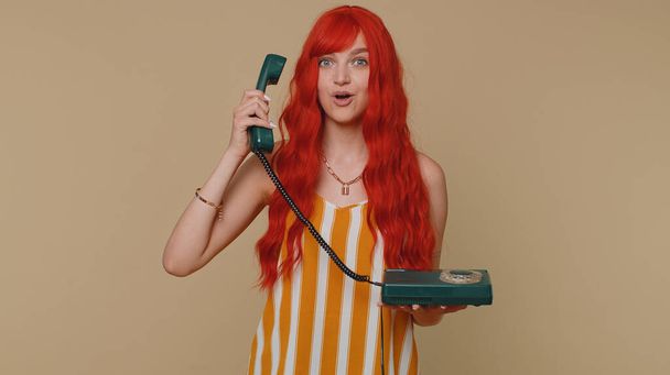 Hey you, call me back. Cheerful one ginger woman secretary in tank top talking on wired vintage telephone of 80s, says hey you call me back. Young redhead girl posing isolated on beige wall background - Photo, Image