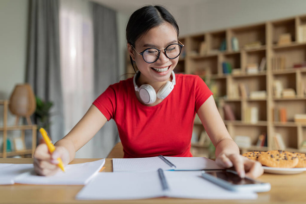 Ευτυχής έφηβος Κινέζα μαθήτρια με γυαλιά σπουδάζουν στο σπίτι, πληκτρολογώντας στο τηλέφωνο στο εσωτερικό του σαλονιού. Home education, social media chat, εργασία, προετοιμασία εξετάσεων και νέα εφαρμογή λόγω πανδημίας covid-19 - Φωτογραφία, εικόνα
