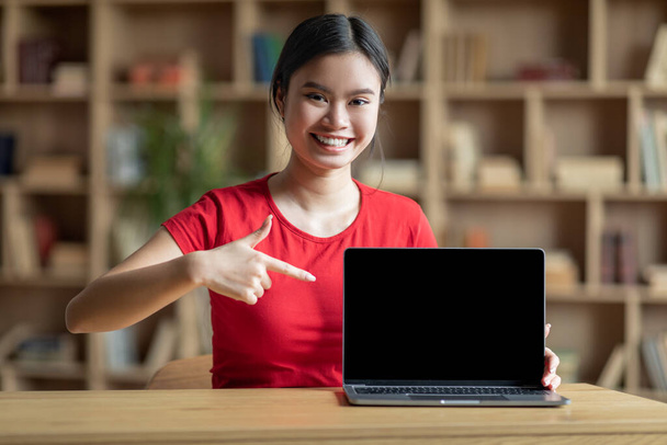 Χαμογελώντας έφηβος κορεάτης κυρία δείχνουν δάχτυλο στον υπολογιστή με κενή οθόνη για μελέτη στο εσωτερικό του δωματίου. Σύγχρονη εκπαίδευση στο σπίτι, εργασία εξ αποστάσεως, συσκευή για online μάθημα και καλή ιστοσελίδα λόγω covid-19 πανδημία - Φωτογραφία, εικόνα