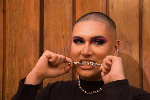 Portrait of non-binary person, young and South American, heavily make up biting a metal chain with teeth, with a wooden wall in the background. Concept queen, lgbtq+, pride, queer. - Photo, image