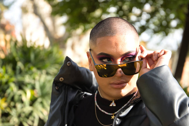 Portrait of non-binary person, young and South American, heavily make up, looking over mirrored sunglasses and surrounded by greenery. Concept queen, lgbtq+, pride, queer. - Foto, imagen