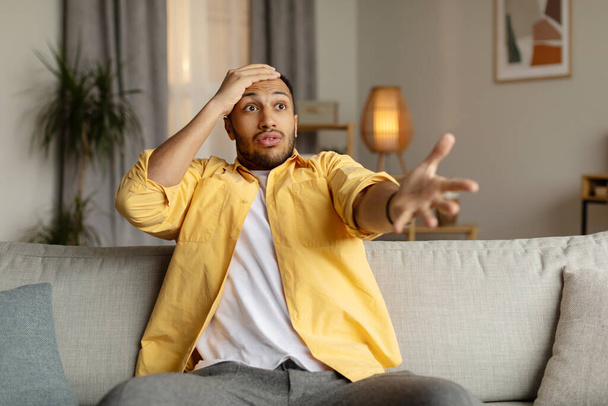 Emotional young black man expressing indignation, shocked by bad news or defeat of sports team on TV at home. Millennial African American guy feeling unpleasantly surprised. Negative human emotions - Photo, Image