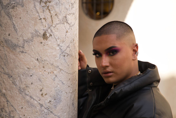 Portrait of non-binary person, young and South American, heavily make up and with serious gesture leaning on a marble column. Concept queen, lgbtq+, pride, queer. - Foto, imagen