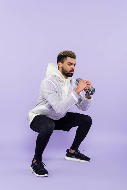 full length of bearded african american sportsman doing squat with dumbbell on purple  - Foto, immagini