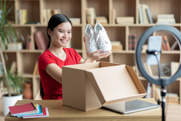 Happy young chinese lady takes out shoes from cardboard box and shoots video blog in living room interior. Shopping online at home, app, delivery service, sale and unpacking due covid-19 quarantine - Photo, Image